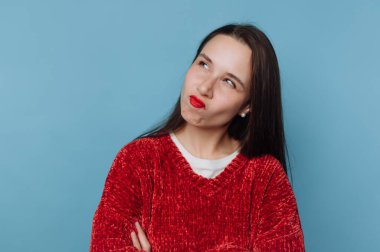 Kırmızı kazaklı kadın, mavi arka planda şaşkın bir ifadeyle düşünüyor..