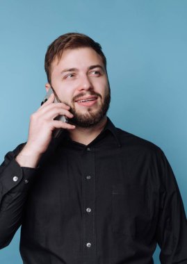 Siyah gömlekli adam mavi arka planda akıllı telefondan konuşuyor..