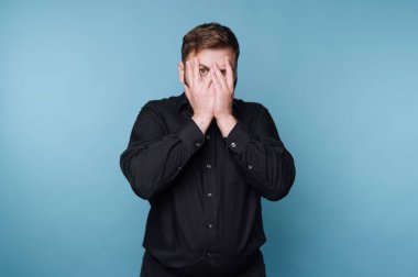 Siyah gömlekli adam yüzünü mavi arka planda kapatıyor..