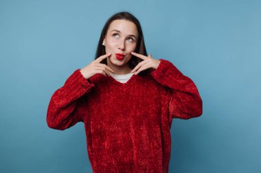 Kırmızı kazaklı kadın mavi arka planda şakacı poz veriyor..