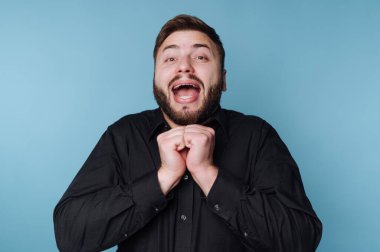 Siyah gömlekli heyecanlı adam mavi arka planda sevinç ifade ediyor..