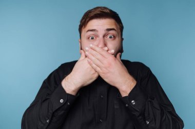 Şaşırmış bir adam iki eliyle mavi bir arka planda ağzını kapatır..