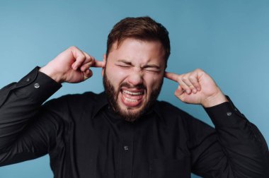 Siyah gömlekli adam kulaklarını mavi arka planda kapatıyor..