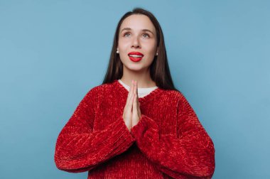 Uzun saçlı, kırmızı kazaklı genç bir kadın ellerini mavi bir arka plana karşı dua pozisyonu alarak kaldırıyor.