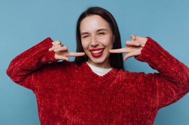 Kırmızı kazaklı, gülümseyen kadın mavi arka planda şakacı poz veriyor..