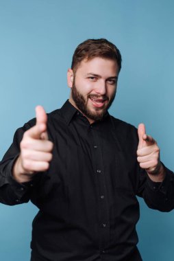 Siyah gömlekli adam mavi arka planda parmaklarını şakayla işaret ediyor..