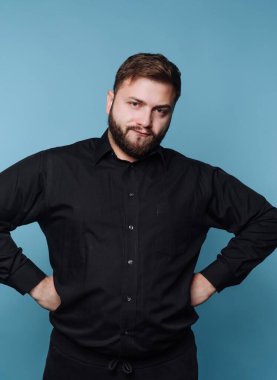 Siyah gömlekli bir adam elleri kalçasında mavi arka planda poz veriyor..