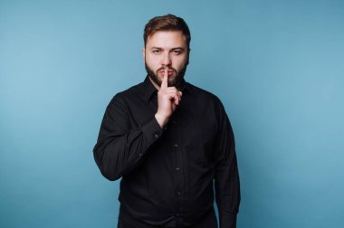 Siyah gömlekli adam mavi arka plana karşı şşşş hareketi yapıyor..