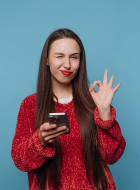 Kırmızı kazaklı kadın göz kırpıyor ve telefonu tutarken tamam işareti gösteriyor..