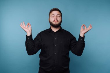 Siyah gömlekli adam, mavi arka planda gözleri kapalı meditasyon yapıyor..