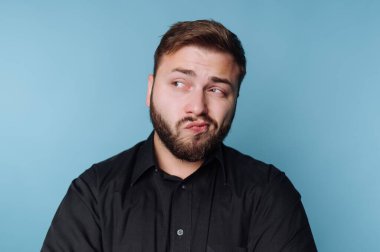 Siyah tişörtlü sakallı bir adam mavi arka planda şüpheli bir ifade takınıyor..