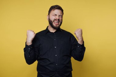 Siyah gömlekli adam sarı arka planda yumruklarını sıkarak kutlama yapıyor..