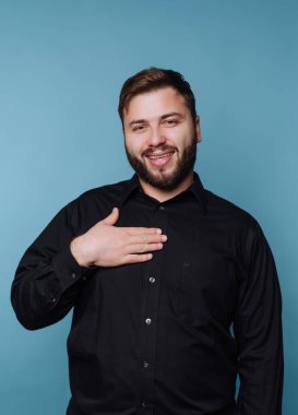 Siyah gömlekli gülümseyen adam mavi arka planda, elleri göğsünde.