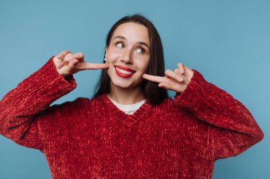 Kırmızı kazaklı kadın gülümsüyor ve yanaklarını gösteriyor.