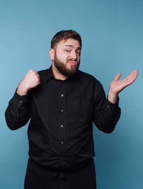 Siyah gömlekli bir adam mavi arka plana karşı karışık bir ifadeyle omuz silkiyor..