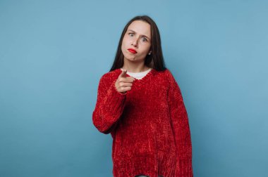 Kırmızı kazaklı kadın mavi arka planda ciddi bir ifadeyle işaret ediyor..