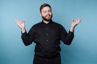 Siyah gömlekli adam, mavi arka planda sakin bir ifadeyle meditasyon yapıyor..