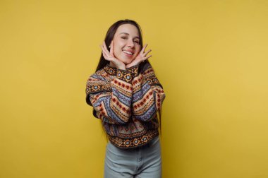 Young woman in patterned sweater posing playfully against yellow background.