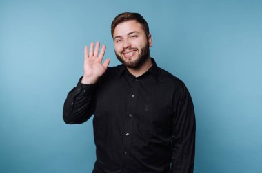 Siyah gömlekli bir adam mavi arka planda el sallıyor..