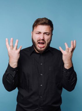 Sinirli bir adam, öfkesini ellerini kaldırarak ifade ediyor, mavi arka planda siyah bir gömlek giyiyor.