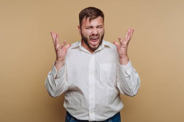 Stüdyoda elini kaldırıp bağıran bir adam var. Yüz ifadesi hayal kırıklığını gösteriyor. Beyaz gömlek ve kot pantolon giyiyor. Arkaplan bej.