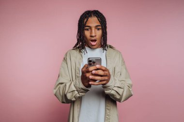 Örgülü saçlı genç bir adam akıllı telefonuna şaşırmış bir ifadeyle bakıyor. Arkaplan pembedir, ve iç mekan ayarları kişinin odaklanmasına katkı sağlar..