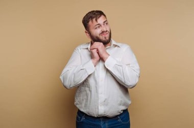 Bir adam elleri birarada durur, gülümser ve yana bakar. Beyaz gömlek ve kot pantolon giyiyor. Arkaplan sade bir açık renk.