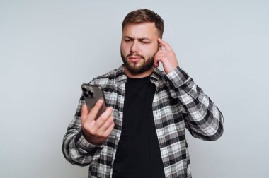 Bir adam içeride durmuş, çatık kaşlı akıllı telefonuna bakıyor. Ekrandaki bir şeyi derinden düşünüyor gibi görünüyor. Eli başının üzerinde duruyor..