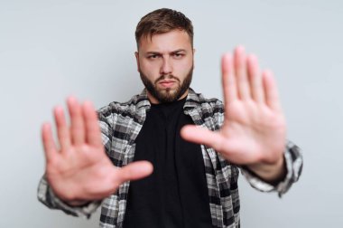 Bir adam düz bir arkaplanın önünde durur ve ellerini kaldırır. Ciddi ciddi bakıyor ve siyah bir gömleğin üzerine kareli bir gömlek giyiyor..