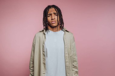 Genç bir insan yumuşak pembe bir arka planın önünde duruyor. Saçları örgülü ve bol gömlek giyiyorlar. Ayarlar basit ve onların ifadesine odaklı..