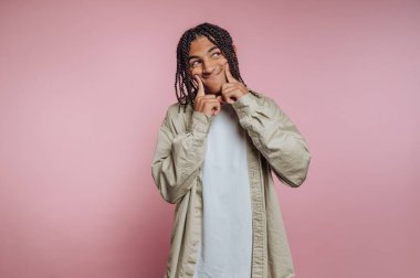 Genç bir insan pembe bir duvara karşı duruyor, parmakları yanaklarında neşeli bir yüz yapıyor. Neşeli görünüyorlar ve açık renkli bir gömlek ve ceket giyiyorlar..