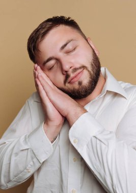 Beyaz gömlekli bir adam gözlerini kapatıyor ve ellerini yüzünün üzerine koyuyor. Stüdyo çekimi için poz verirken hafifçe gülümsüyor..