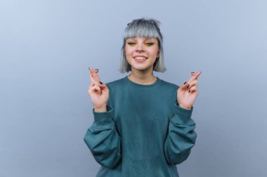 Genç bir insan gri bir arkaplanda duruyor, gülümsüyor ve şans diliyor. Basit, uzun kollu bir bluz giyiyorlar ve açık renk saçları var..