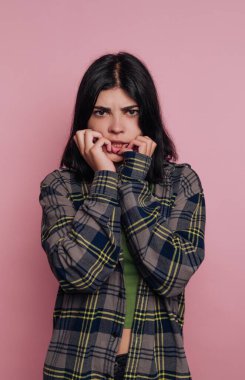 Genç bir insan pembe bir arka fonun önünde duruyor, tırnak yerken endişeli bir yüz ifadesi sergiliyor. Kişi yeşil bir bluzun üzerine ekose gömlek giyer ve doğrudan izleyiciye bakar..