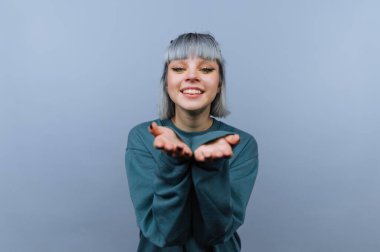 Genç bir kadın gülümseyerek sade bir arka planın önünde duruyor. O, dostça bir jest yapmak için ellerini onun önünde uzattı. Gri saçları ışıkta parlıyor..