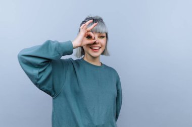 Genç bir insan açık gri bir arka plana karşı duruyor ve elleriyle bir çember oluştururken gülümsüyor. Ayarlar dikkat dağıtıcı olmadan parlak ve basit görünüyor..