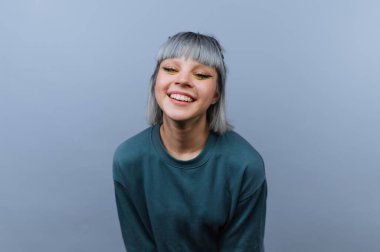 Genç bir kadın yüzünde büyük bir gülümsemeyle oturuyor. Kısa gri saçları var ve yeşil bir gömlek giyiyor. Arka plan gridir, iyi ışıklandırılmıştır. Özellikleri göze çarpar..