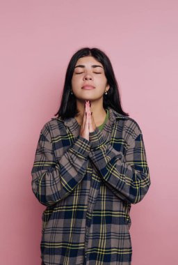 Bir kadın dua ederken ellerini birbirine bastırır. Gözleri kapalı ve ekose bir gömlek giyiyor. Arka plan pembe ve sahne bir yansıma anını akla getiriyor..