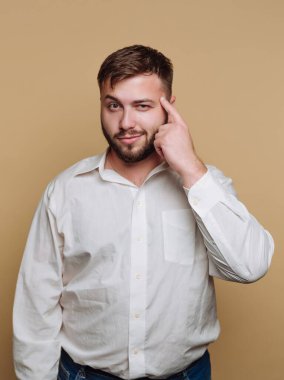 Bir adam sade bir arka planın önünde duruyor ve hafifçe gülümserken tek eliyle kafasını işaret ediyor. Düşüncelere dalmış ve sohbete dalmış gibi görünüyor..