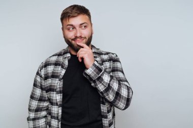 Genç bir adam kapalı bir alanda duruyor. O, kareli bir gömlek ve siyah bir gömlek giyiyor. Parmağı çenesinde düşünceli görünüyor..