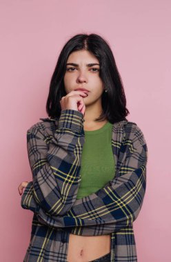 Genç bir kadın kollarını kavuşturmuş pembe bir duvarın önünde duruyor. Dudağını ısırırken düşünceli görünüyor. Yeşil bir bluz ve ekose bir gömlek giyiyor. Sıradan bir tarzı var..