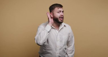 Beyaz gömlekli bir adam içeride duruyor. Şaşırmış bir ifadesi var ve sanki daha iyi bir şey duymaya çalışıyormuş gibi elini kulağına dayıyor..