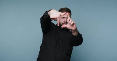 Bir adam mavi bir arka planın önünde duruyor. Elleriyle kare bir çerçeve yapıyor. Poz verirken özgüven gösteriyor. Onun siyah gömleği arka planla çelişiyor..