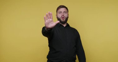 Siyah gömlekli bir adam önemli bilgileri açıklamak için el işaretleri kullanır. Sarı bir arka planın önünde duruyor, konuşurken seyirciyi kendine çekiyor..