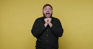 Sakallı biri parlak sarı bir arka planın önünde duruyor. Komik suratlar yapıyor ve jestler ve kahkahalarla heyecan gösteriyor. Anın tadını çıkarıyor gibi..