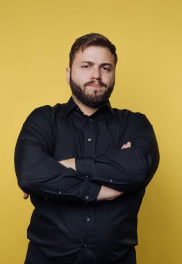 Siyah gömlekli, kendine güvenen, sarı arka planda kolları bağlı bir adam..