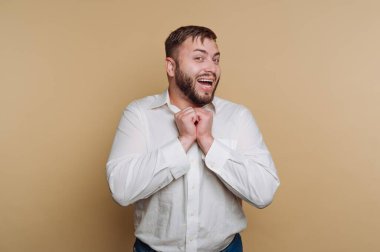 Beyaz tişörtlü, elleri bağlı, bej arka planda gülümseyen adam.