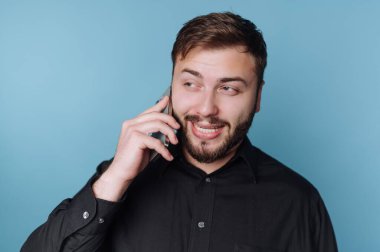 Siyah gömlekli adam mavi arka planda akıllı telefondan konuşuyor..