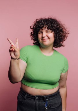 Yeşil tişörtlü, kıvırcık saçlı, gülümseyen kişi barış işareti yapıyor..