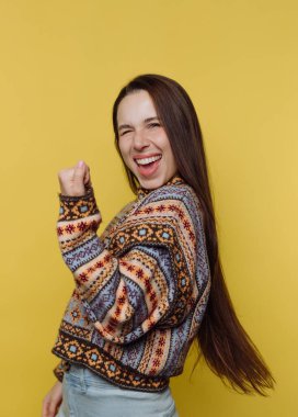 Renkli bir kazak giyen neşeli bir kadın sarı bir arka plana yumruk sallayarak kutluyor..